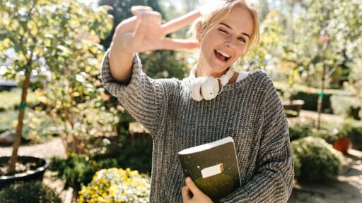 woman in high spirits smiles winks and shows sign of peace Snapshot of girl with notebook posing in botanical garden- living well