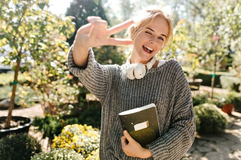 woman in high spirits smiles winks and shows sign of peace Snapshot of girl with notebook posing in botanical garden- living well